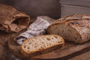Freshly baked sourdough bread loaf and sliced piece on a rustic wooden cutting board with a kitchen towel and paper bag in the background