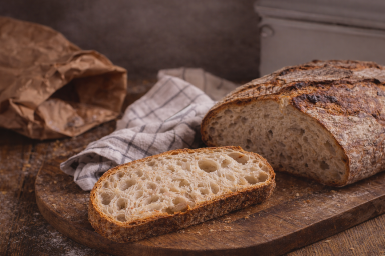 Freshly baked sourdough bread loaf and sliced piece on a rustic wooden cutting board with a kitchen towel and paper bag in the background